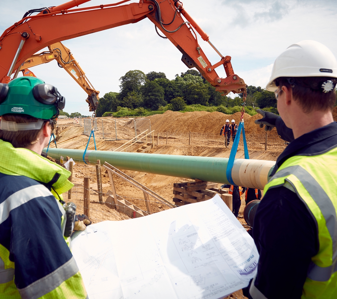 Two construction workers holding works plans and looking out at two cranes on a building site lifting a large pipe