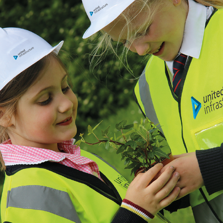 Two children wearing United Infrastructure hard hats and high vis jackets holding a plant